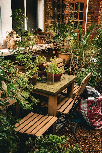 A small garden area adjacent to a brick house features a weathered wooden table surrounded by matching wooden chairs with slatted seats. The table is decorated with various potted plants, including sm