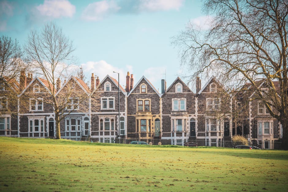A row of Victorian-style terrace houses with dark brick facades, large sash windows, and pitched roofs, situated behind a well-maintained lawn. The lawn features freshly cut, lush green grass with a u